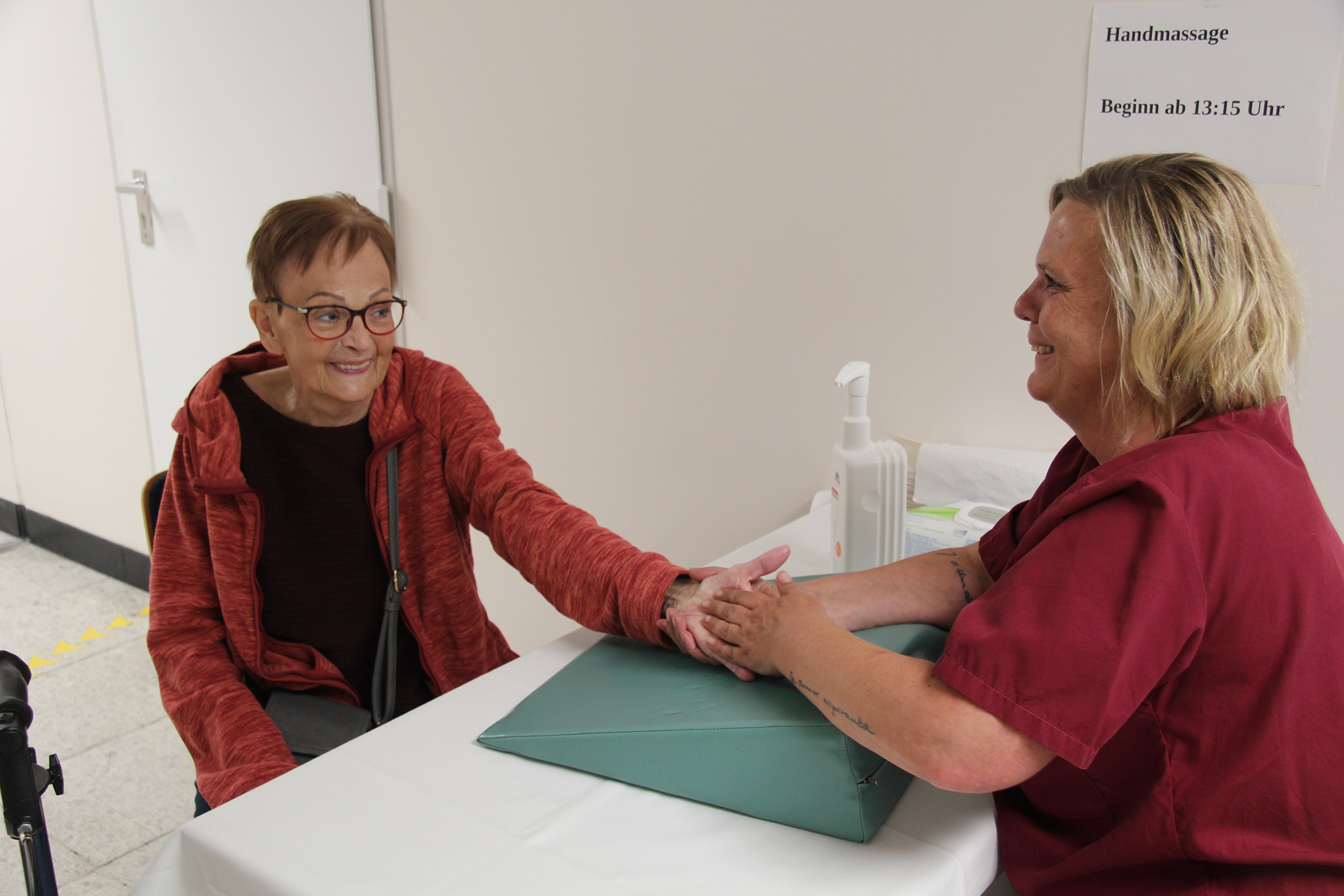 Eine Frau erhält eine Handmassage von einem Mitarbeiter, der an einem Tisch sitzt, beide lächeln und sind sehr freundlich.