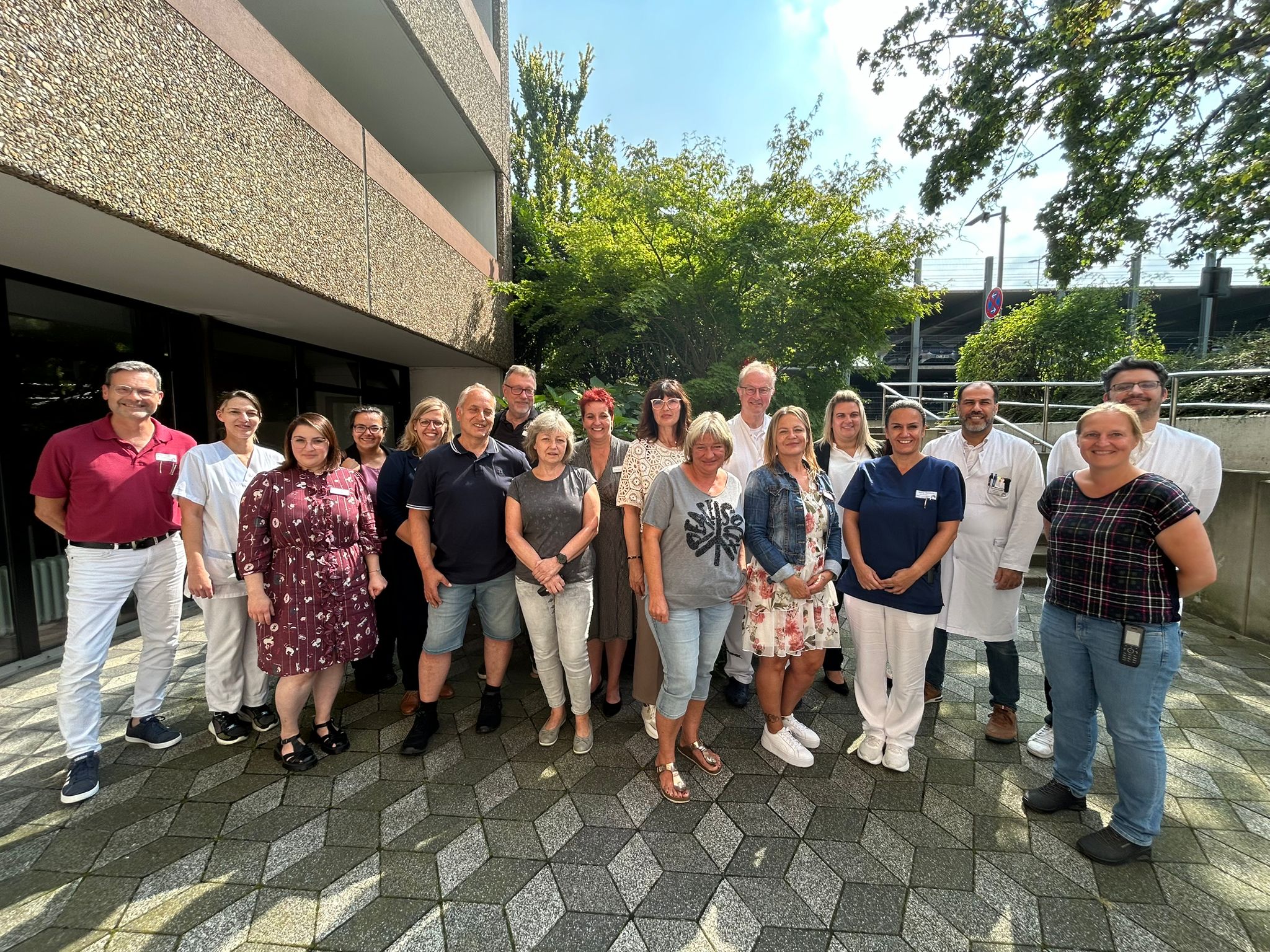 Eine Gruppe Erwachsener steht an einem sonnigen Tag auf einem gepflasterten Platz für ein Gruppenfoto zusammen.