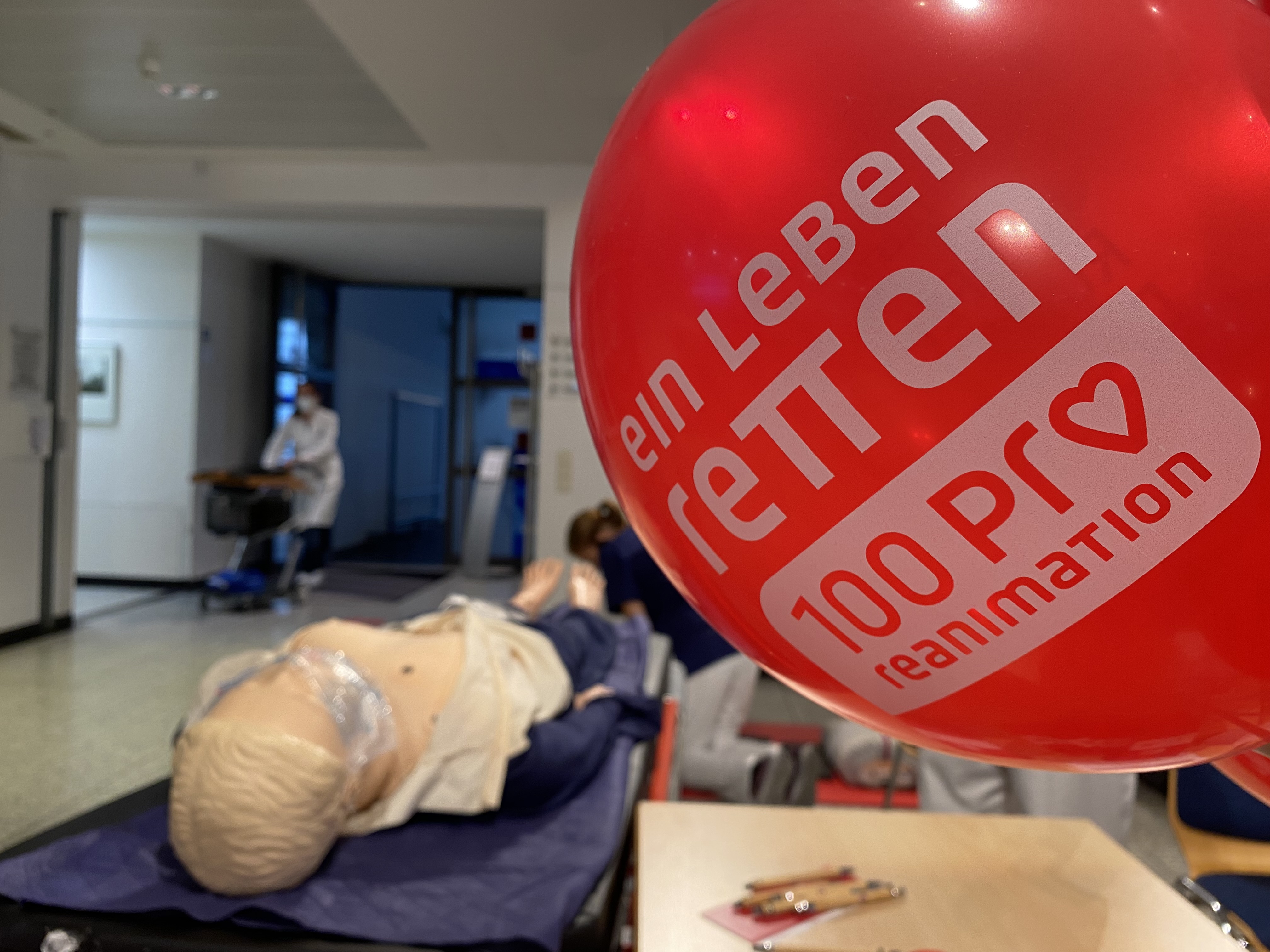 Roter Ballon mit deutschem Text in der Nähe einer HLW-Puppe; Krankenhausflur und Personal im Hintergrund.