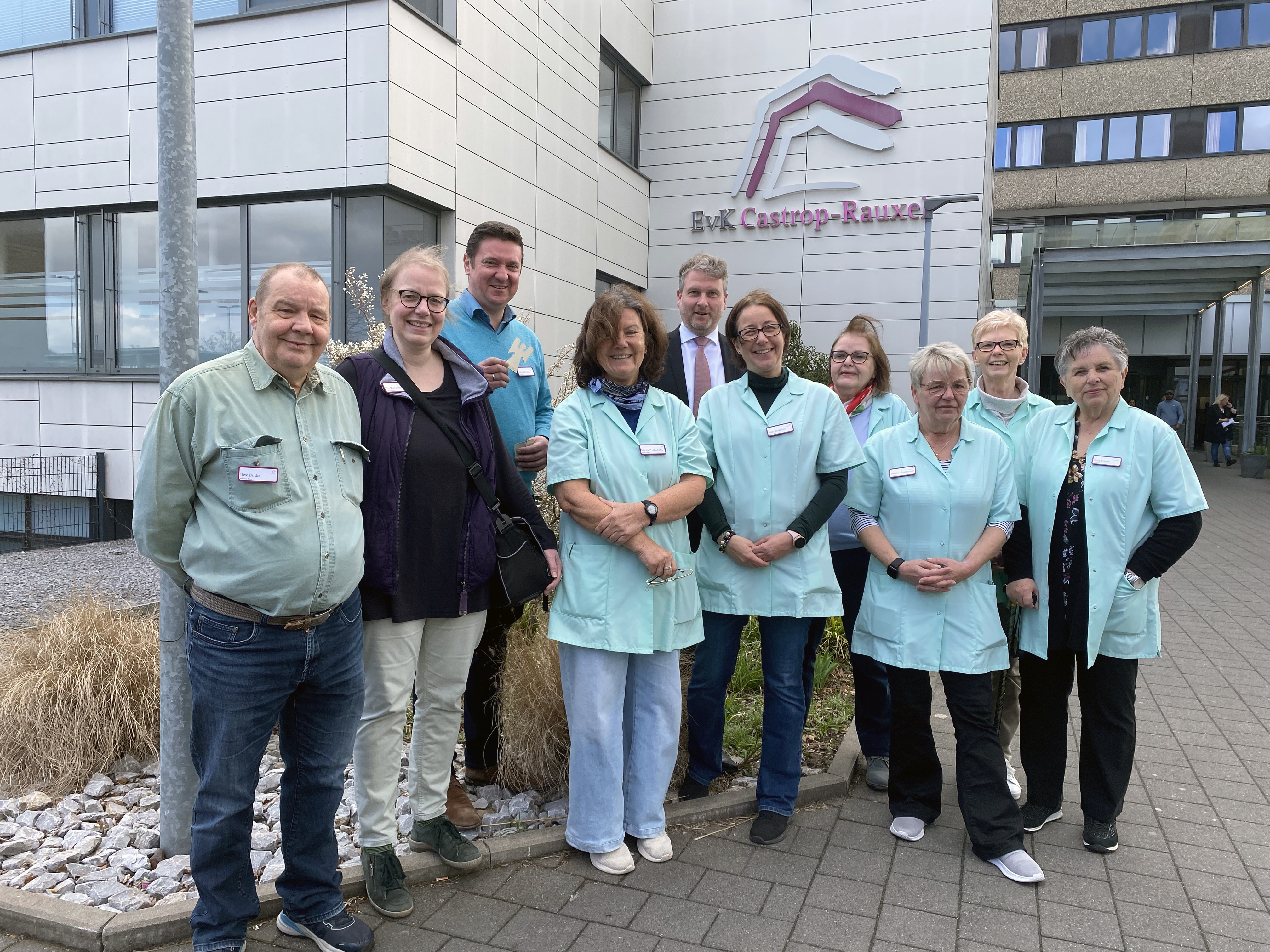 Eine Gruppe von Menschen, einige in hellgrünen Uniformen, steht lächelnd vor einem Gebäude mit einem "BKK"-Schild.