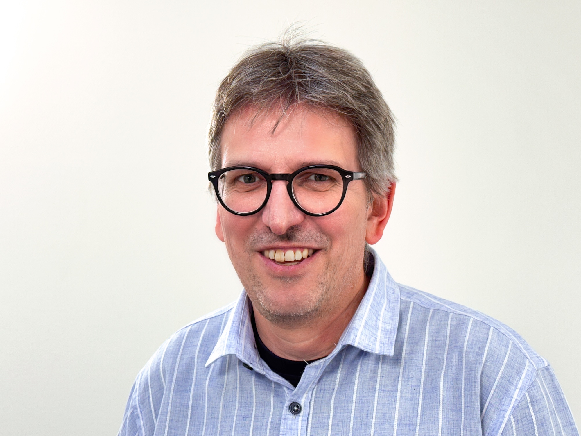 Lächelnder Mann mit Brille und grauem Haar, der ein hellblau gestreiftes Hemd trägt und vor einem schlichten Hintergrund steht.