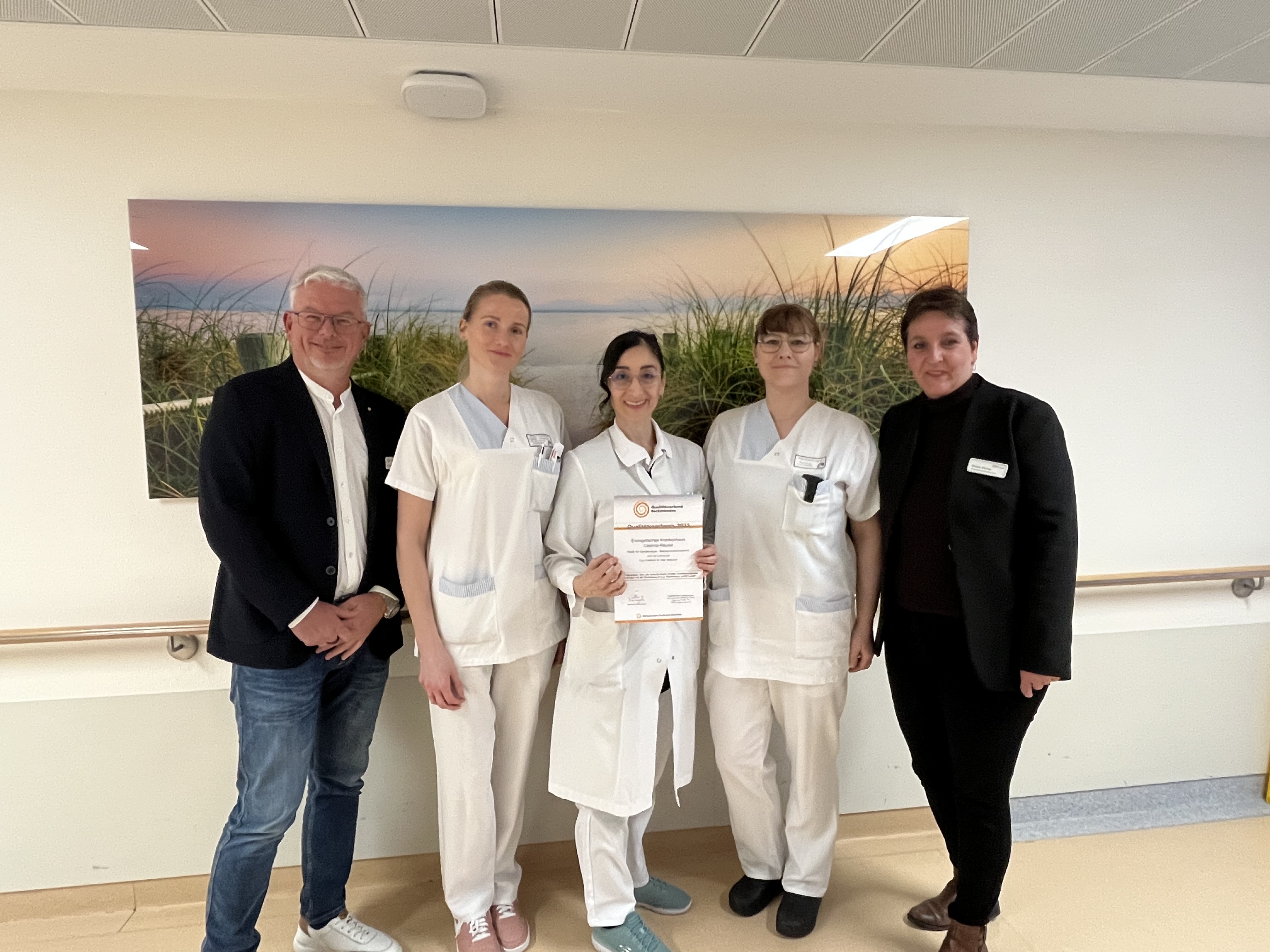 Fünf Personen, darunter drei in weißen medizinischen Uniformen, stehen in einem Innenraum zusammen; eine von ihnen hält eine Urkunde in der Hand.