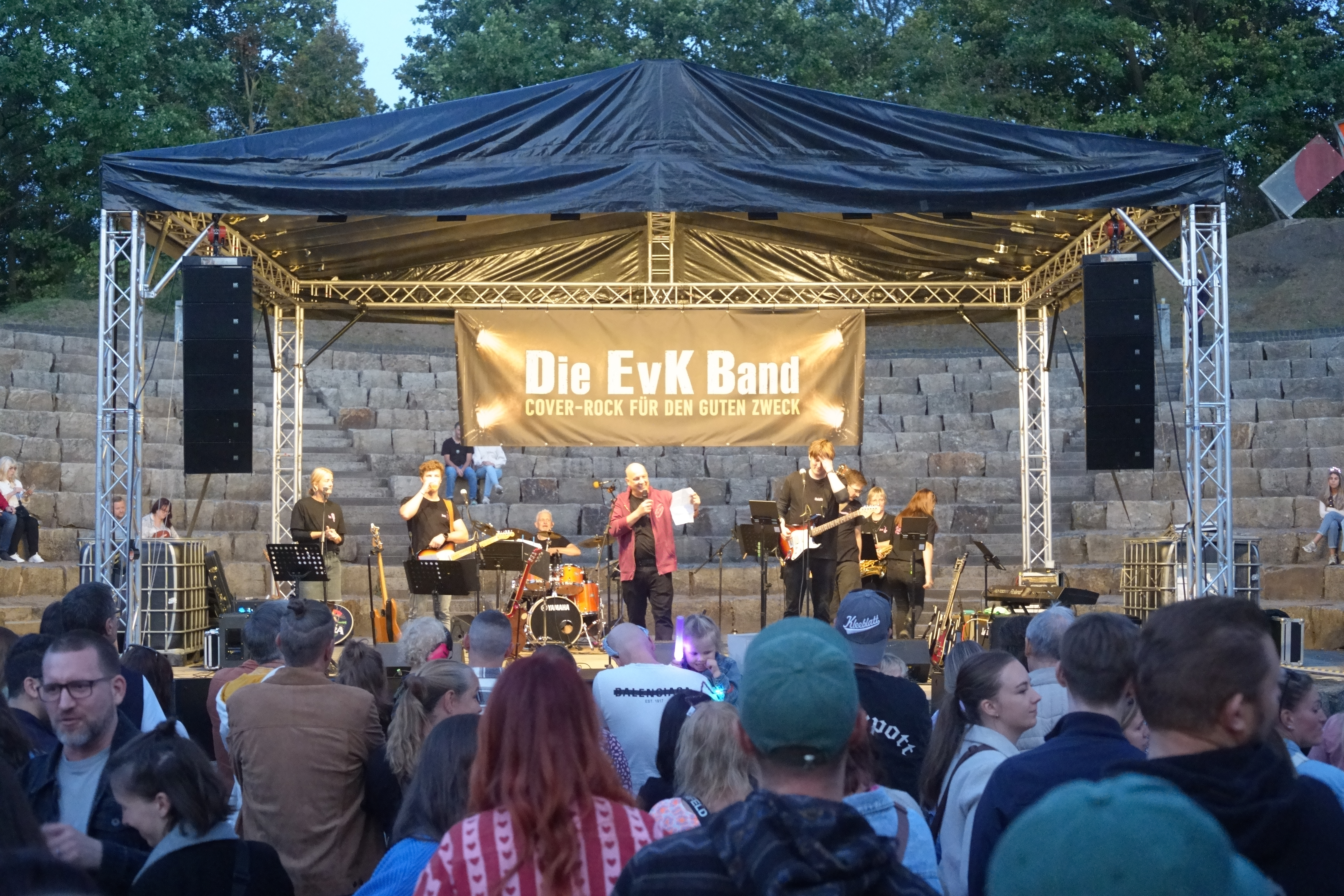Mehrere Musiker stehen auf einer Bühne. Davor stehen einige Menschen. Auf einem Banner steht "Die EvK Band".
