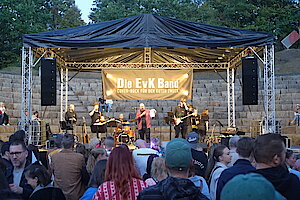 Mehrere Musiker stehen auf einer Bühne. Davor stehen einige Menschen. Auf einem Banner steht "Die EvK Band".