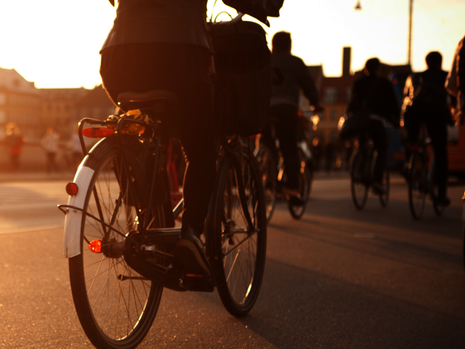 Eine Gruppe von Menschen auf Fahrrädern auf einer Straße in der Stadt bei Sonnenuntergang, wobei das warme Sonnenlicht lange Schatten wirft.