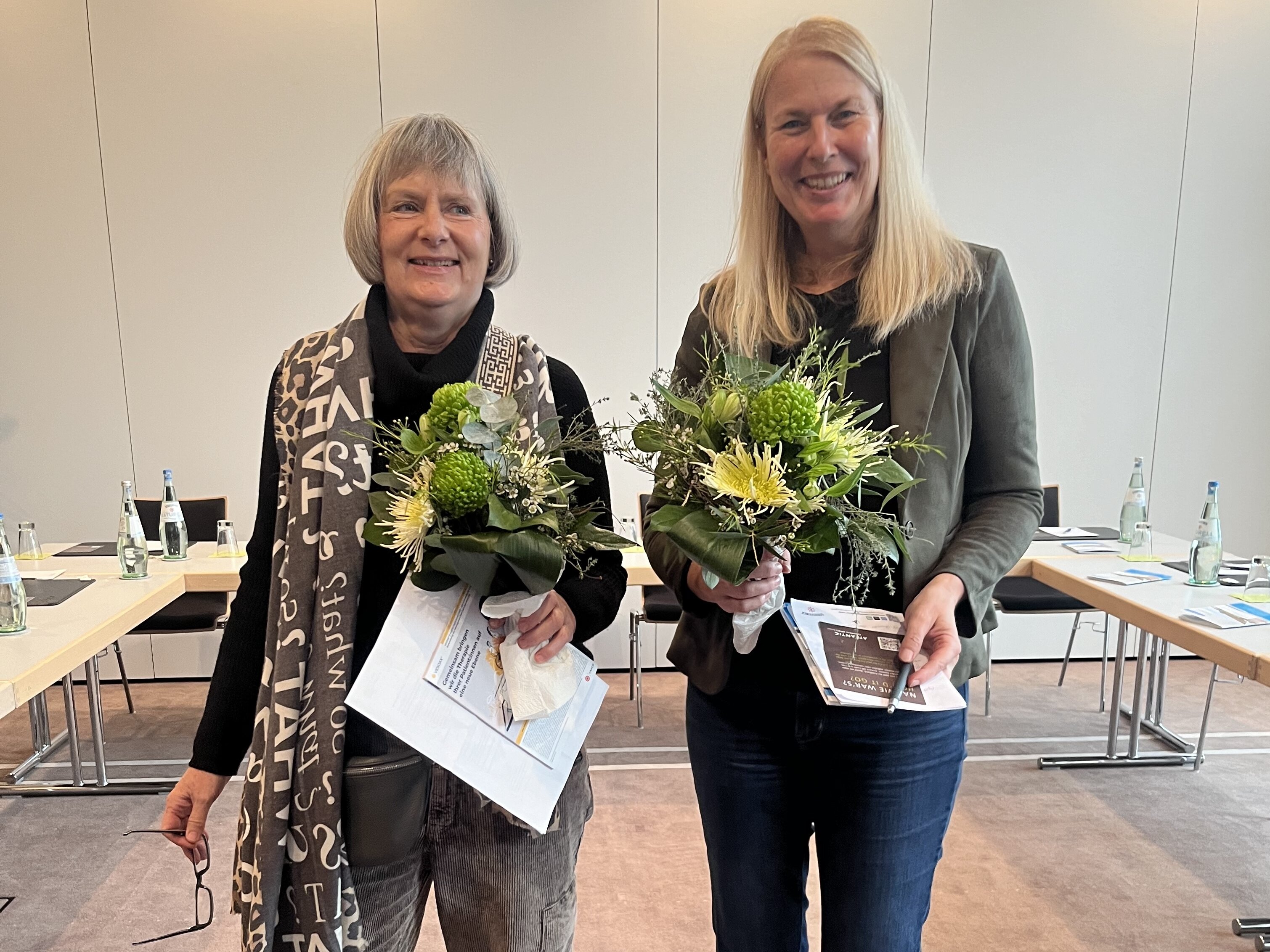 Zwei Frauen stehen lächelnd in einem Haus, jede hält einen Blumenstrauß und Papiere in der Hand, im Hintergrund stehen Tische.