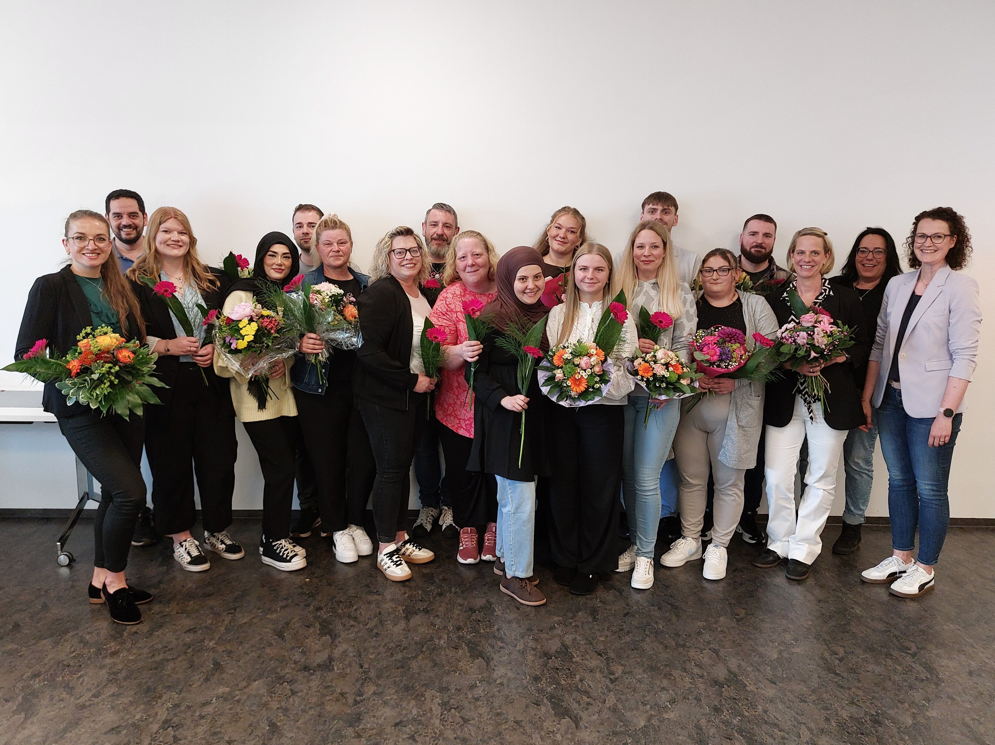 Wundexperten 2026 Junge Menschen, teilweise mit Blumen in den Händen, stehen in mehreren Reihen nebeneinander vor einer weißen Wand und lächeln.