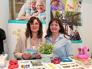 Zwei Frauen stehen hinter einem Informationsstand mit Broschüren und Dekorationen und lächeln in die Kamera.