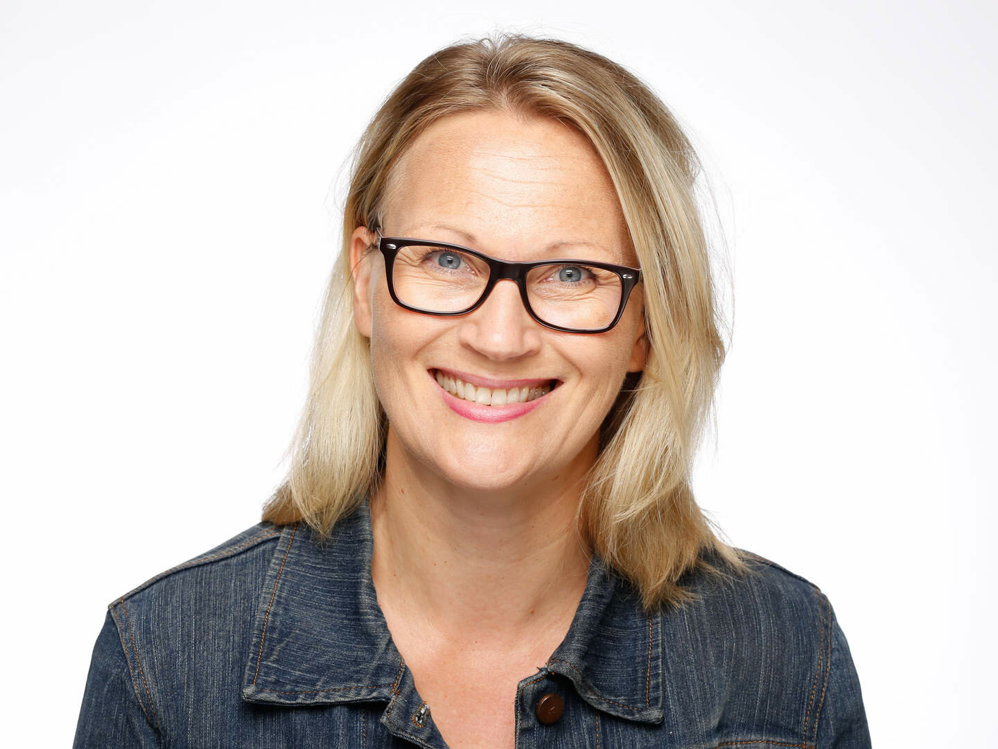 Lächelnde Frau mit blondem Haar, schwarzer Brille und Jeansjacke, vor weißem Hintergrund.