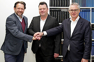 Drei Männer in Anzügen stehen lächelnd und mit erhobenem Daumen vor mit Büchern gefüllten Bücherregalen.