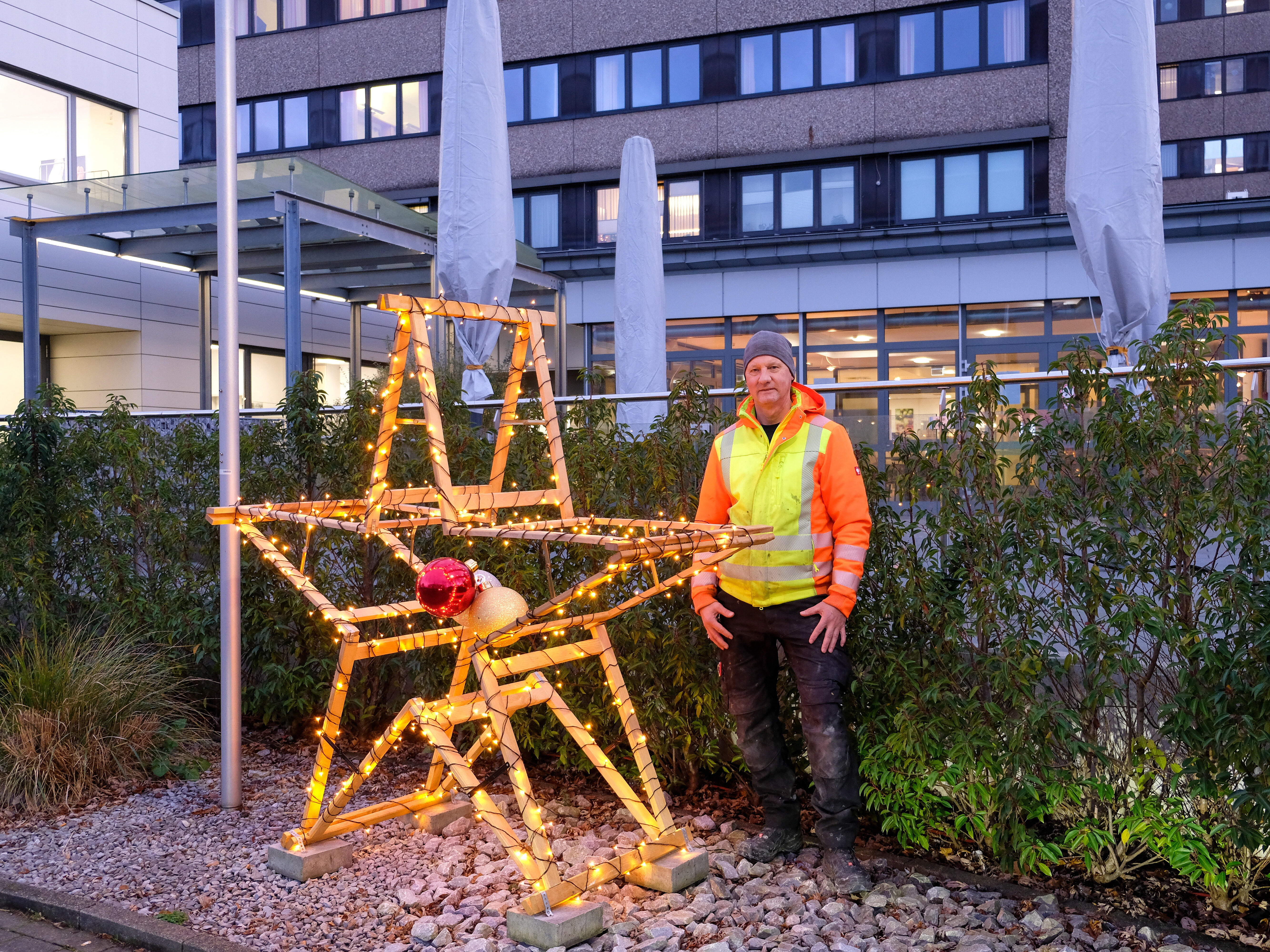 Ein Arbeiter in einer gelben Sicherheitsweste steht neben einer großen beleuchteten Sterndekoration im Freien in der Nähe eines Gebäudes.