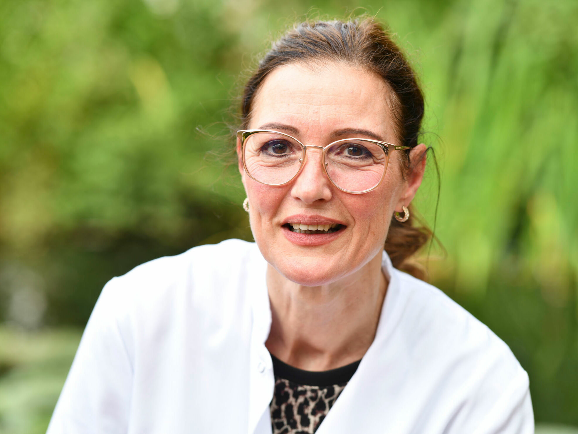 Eine Frau mit Brille und weißem Mantel lächelt im Freien, vor einem grünen, unscharfen Hintergrund.
